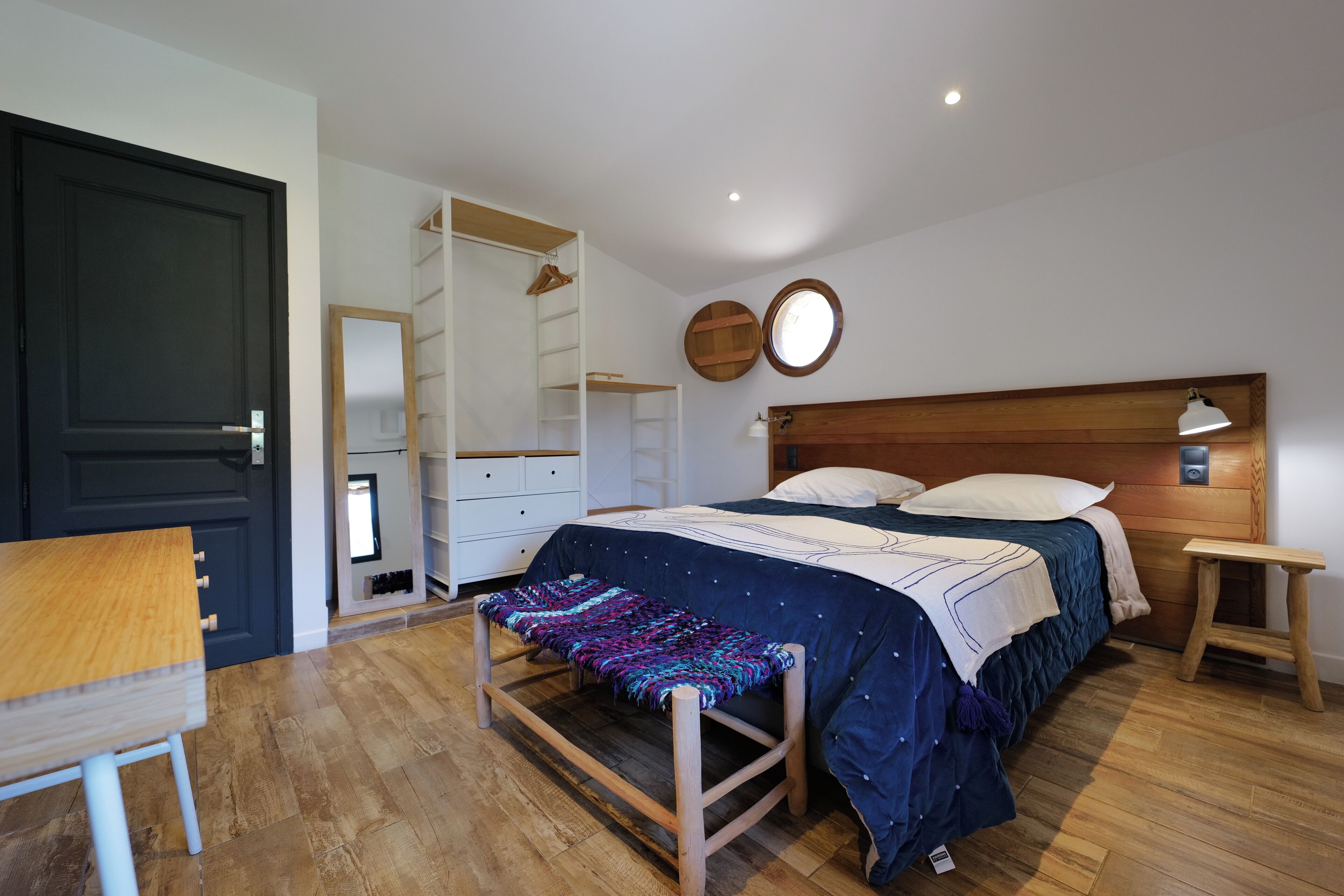 A bedroom with a bed, bedside table, bench, wardrobe, and a mirror on the wall.