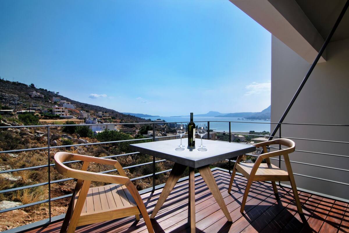 Een balkon met een tafel en twee stoelen, met uitzicht op een mooi landschap met heuvels en een waterpartij.