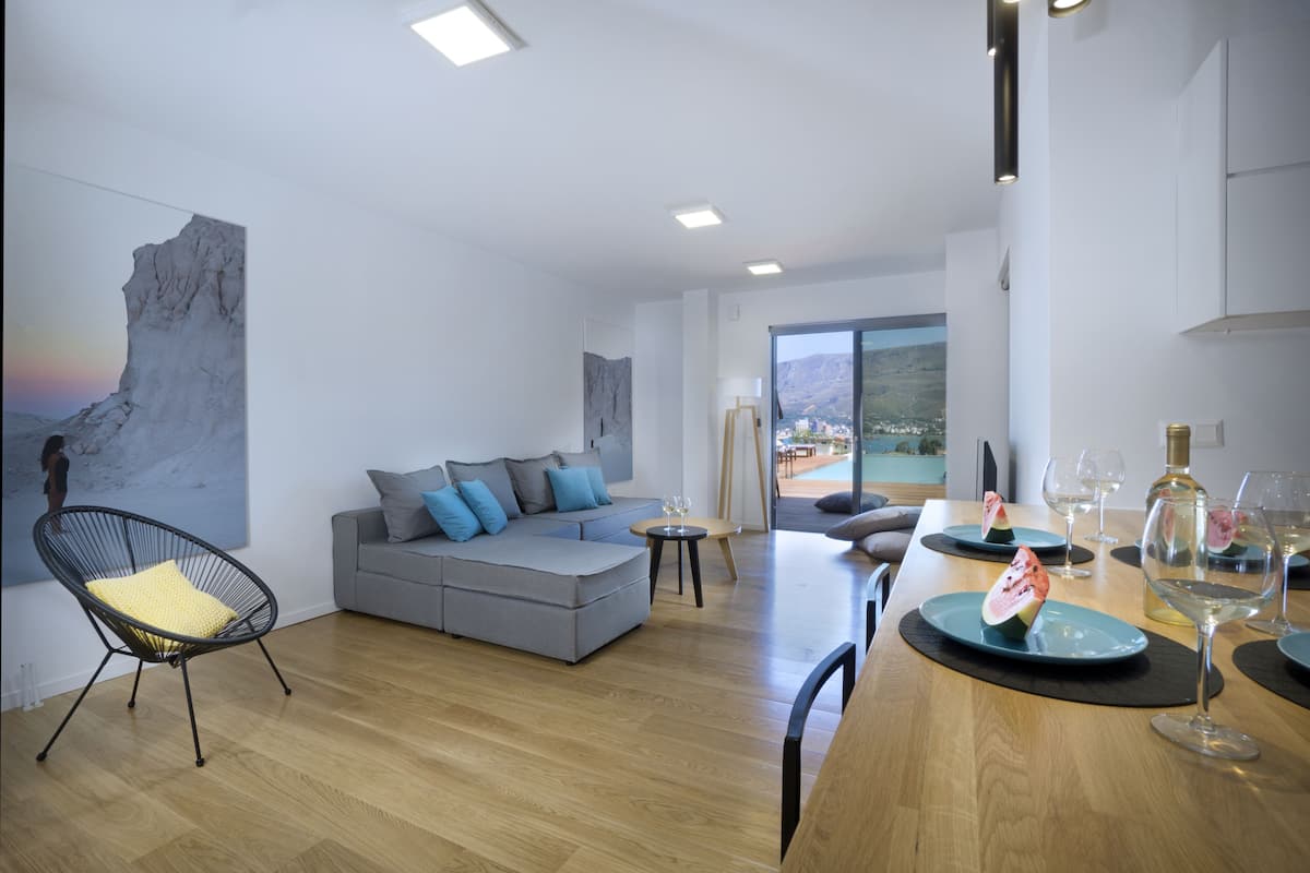Een moderne woonkamer met een grijze modulaire bank, een zwarte frame stoel, een eettafel met borden en glazen, en een groot raam met uitzicht op een bergachtig landschap.