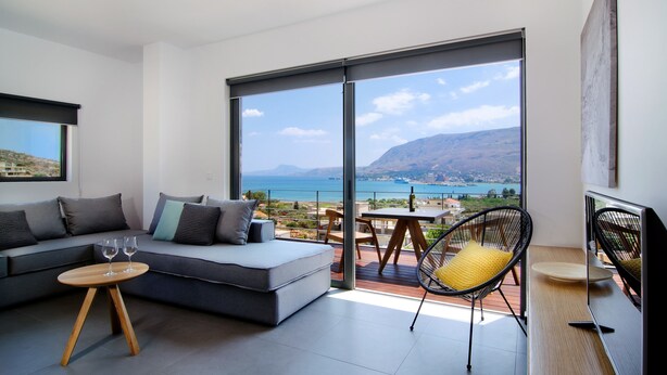 Een moderne woonkamer met een bank, een klein houten tafeltje en een stoel, met uitzicht op een prachtig waterlandschap en heuvels.