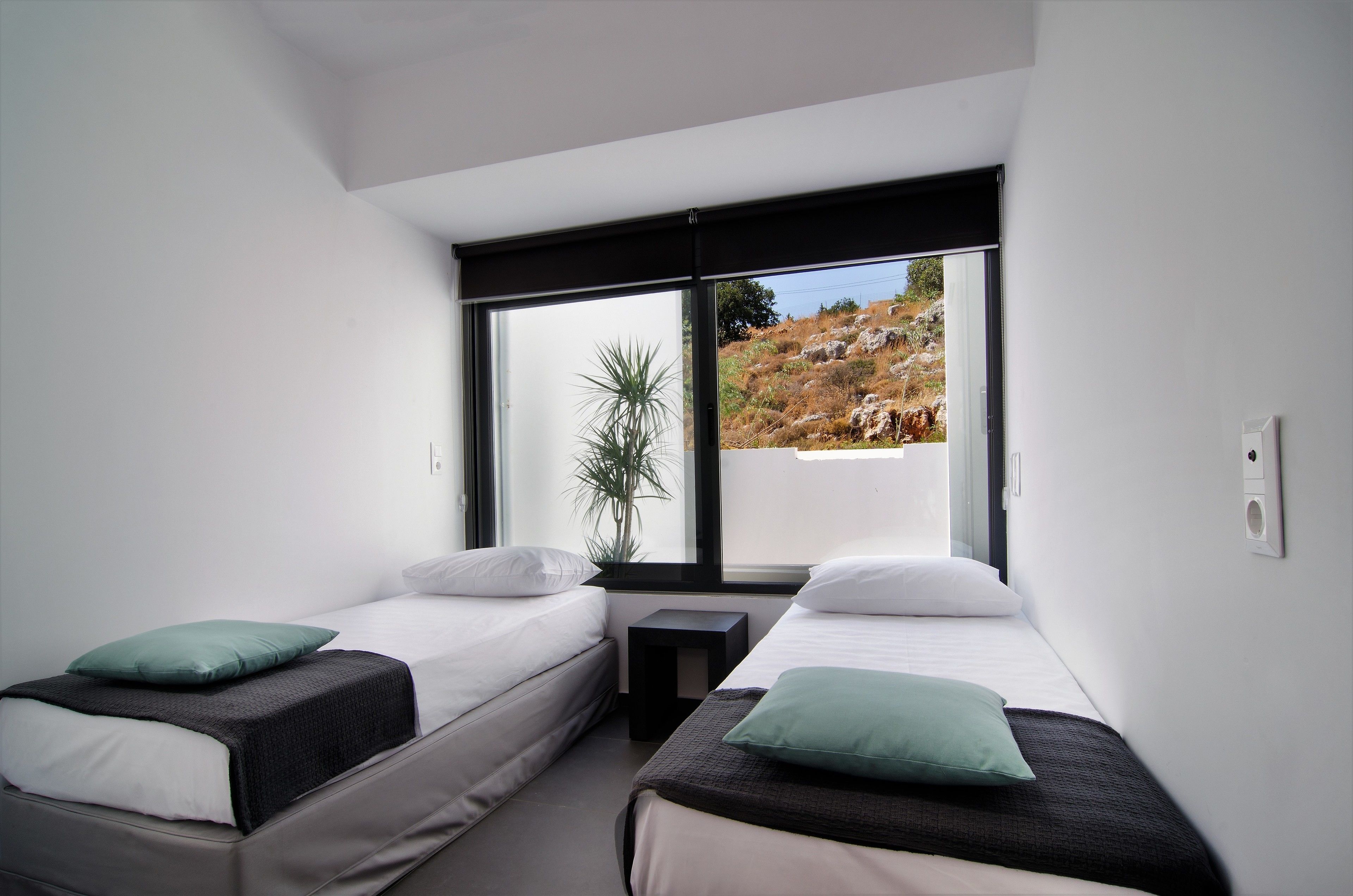 Un dormitorio moderno con dos camas individuales, una mesita de noche pequeña y un ventanal con vistas a un paisaje rocoso.