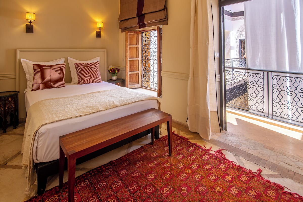A bedroom with a large bed, a bench, a patterned rug, and a balcony with a wrought iron railing.