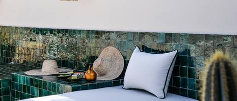 A sunlit room with a green-tiled wall, a white sofa with a cushion, a woven hat, and a small pot on a ledge.