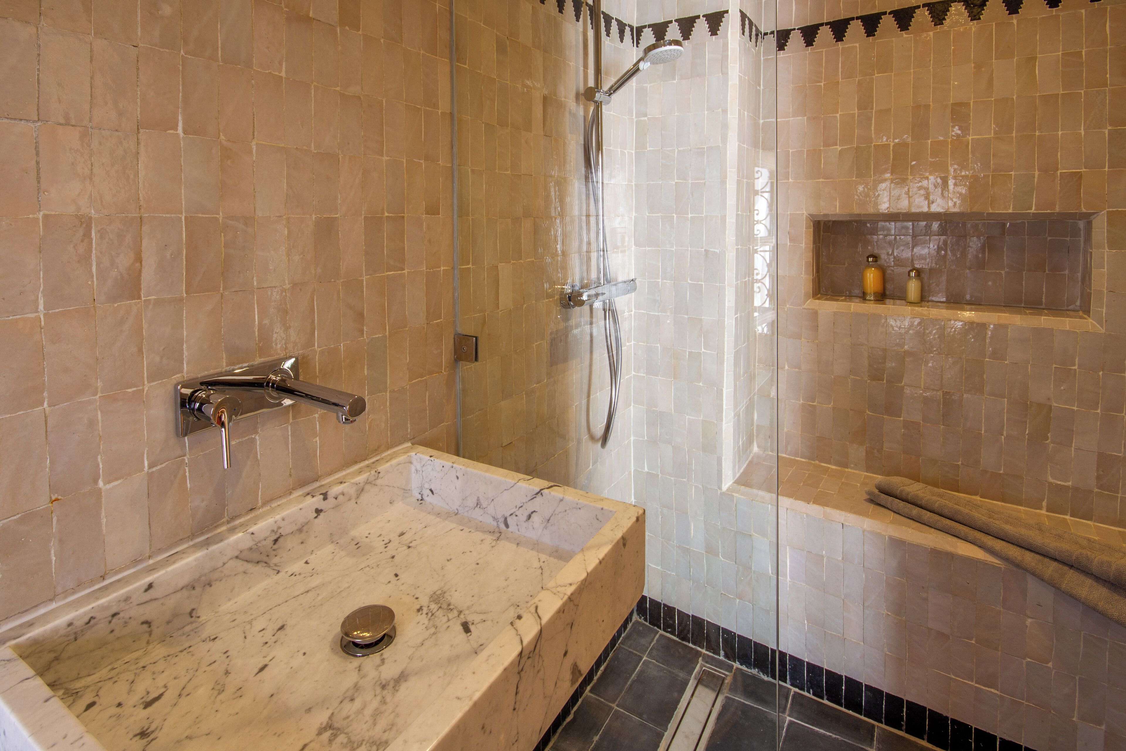 Un baño con lavamanos de mármol, ducha y repisa con artículos de aseo.