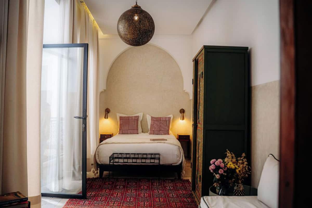 A bedroom with a bed, bedside lamps, a hanging light fixture, a patterned rug, and a door leading to another room.