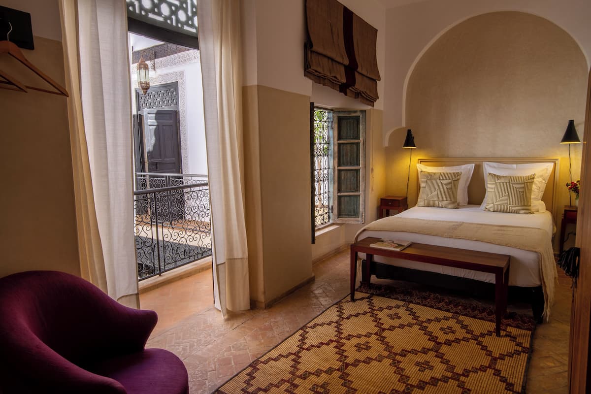 A bedroom with a bed, bedside lamps, a bench, a patterned rug, a chair, and a view of the outside balcony.