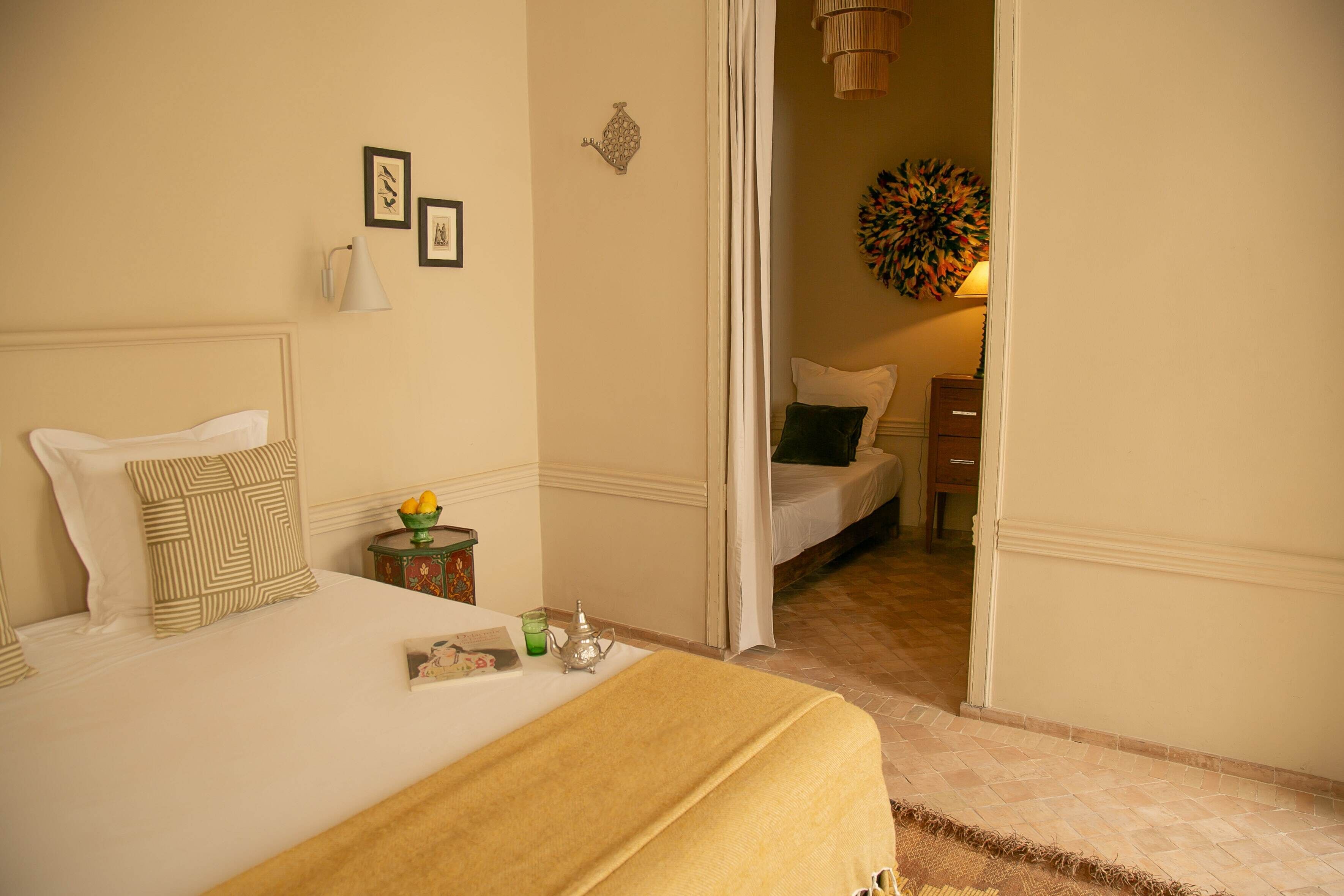 A hotel room with a bed, a nightstand with a lamp, a small table with a book and a teapot, and a door leading to another room.