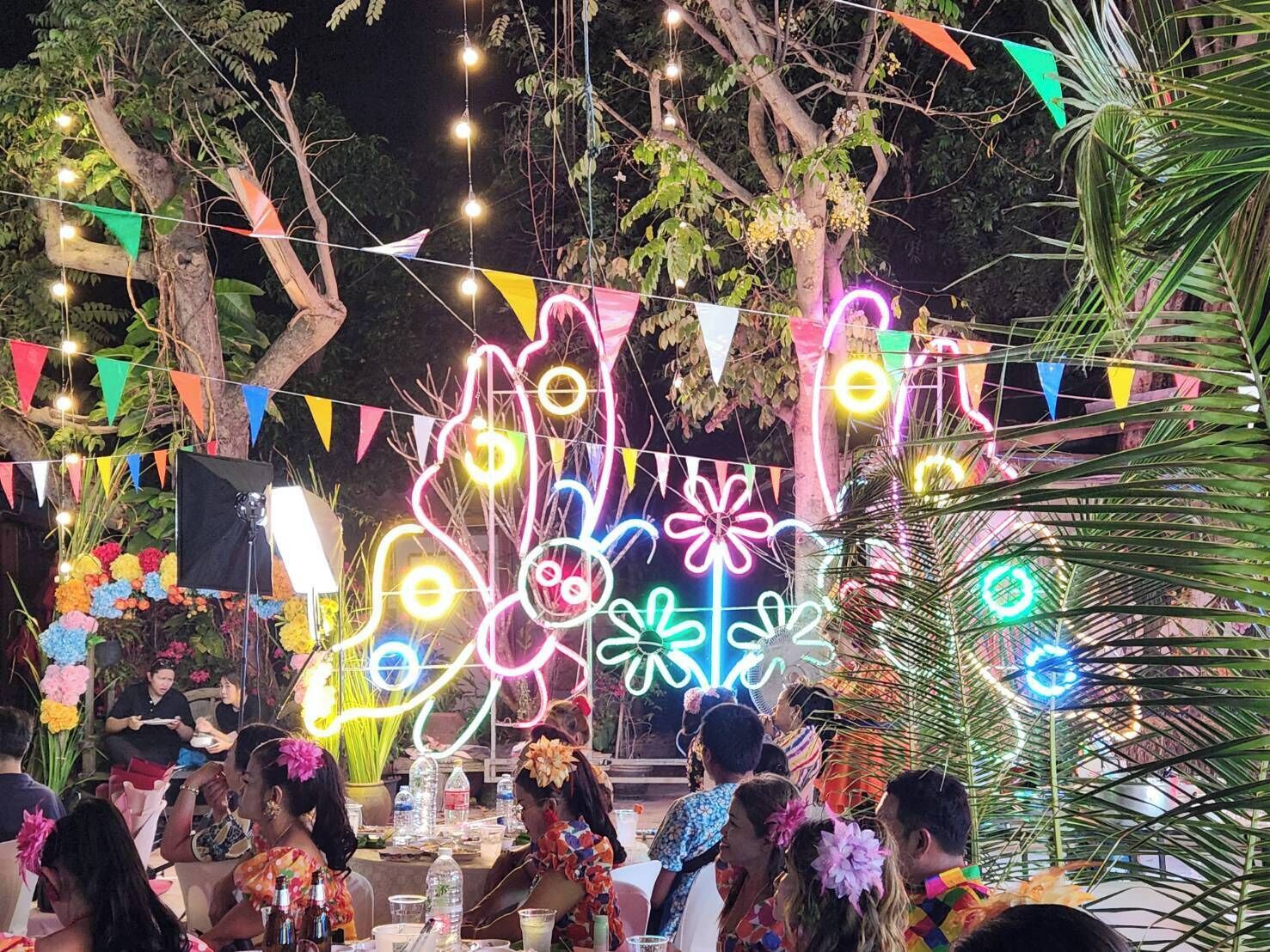 An outdoor event with people seated at tables, decorated with colorful bunting and neon lights.