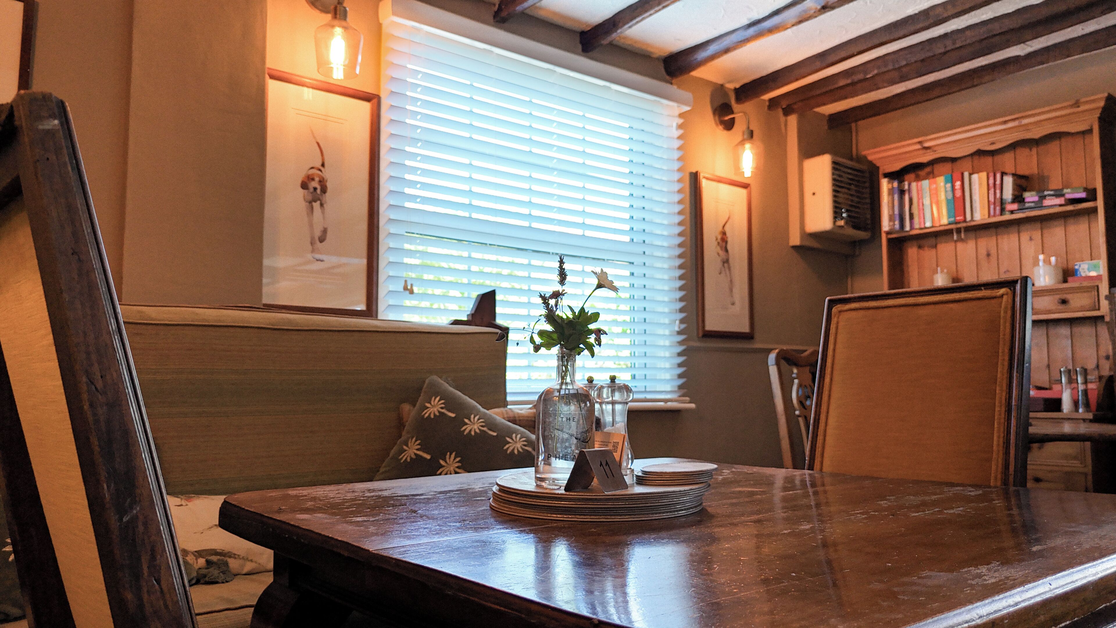 A dining area with a wooden table, a vase with flowers, a window with blinds, and a bookshelf with books.