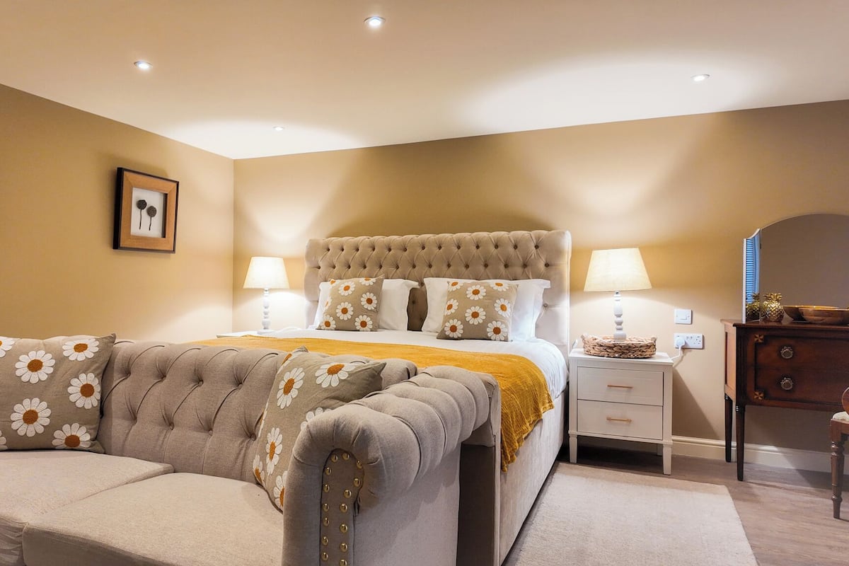 A bedroom with a tufted headboard, a sofa with floral cushions, a bedside table with a lamp, and a dresser.