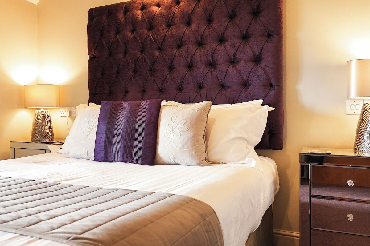 A neatly made bed with a tufted headboard, a bedside table with a lamp, and a nightstand with a decorative item.