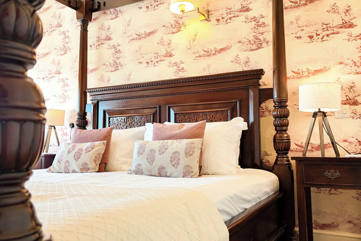 A four-poster bed with a wooden headboard, multiple pillows, and a bedside table with a lamp.