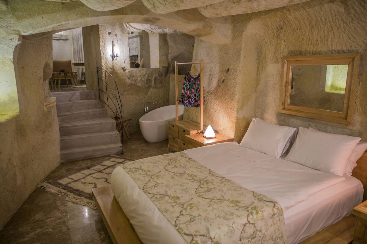 Un dormitorio con cama, mesita de noche, espejo, una silla de madera y baño con bañera e inodoro.