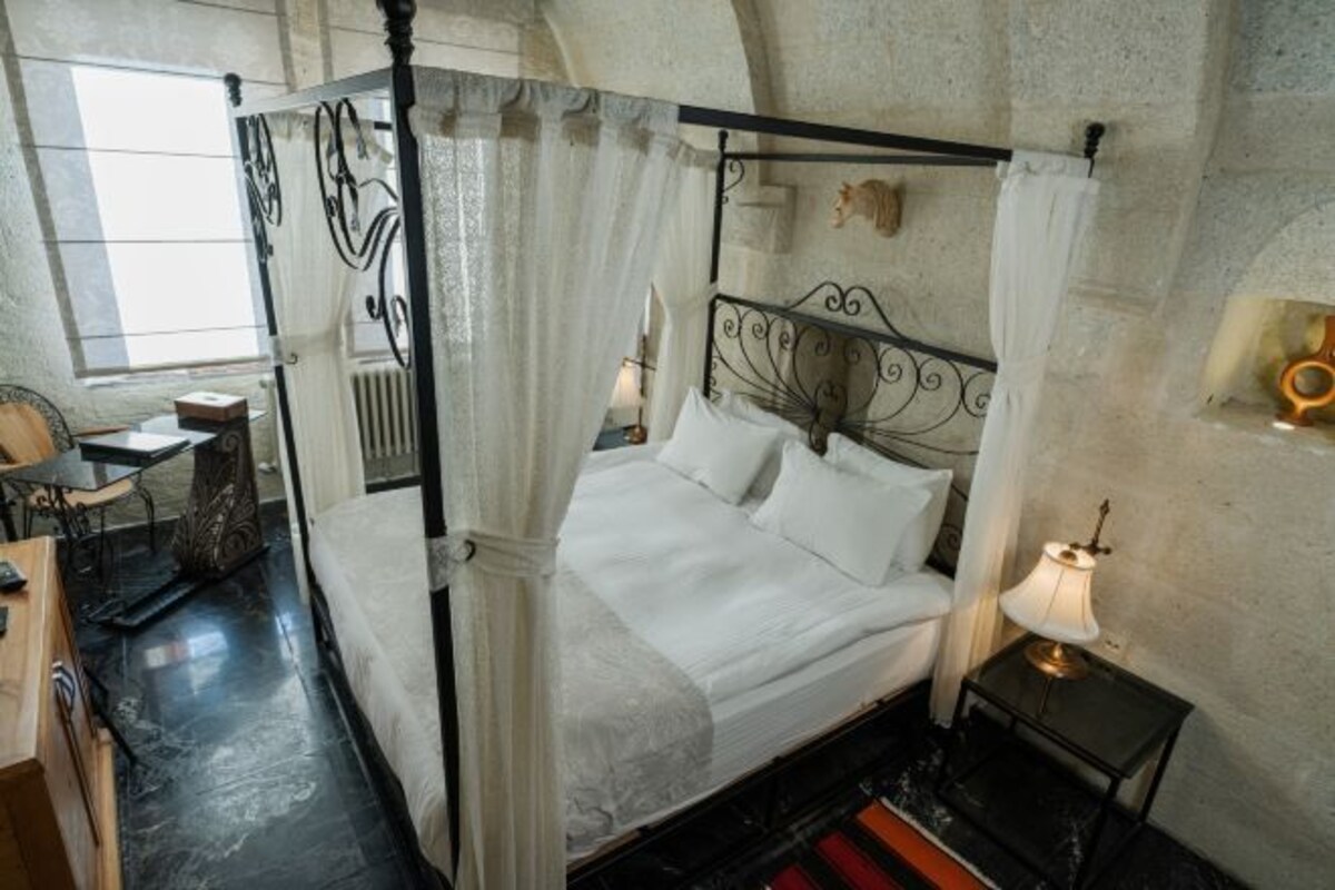 A four-poster canopy bed with a headboard, a bedside table with a lamp, and a small shelf with decorative items.