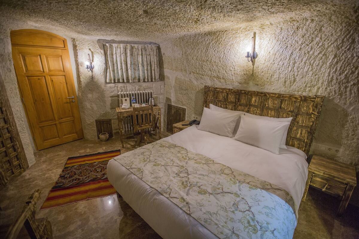 Un dormitorio con puerta de madera, cama con ropa blanca, alfombra estampada y pared de piedra.