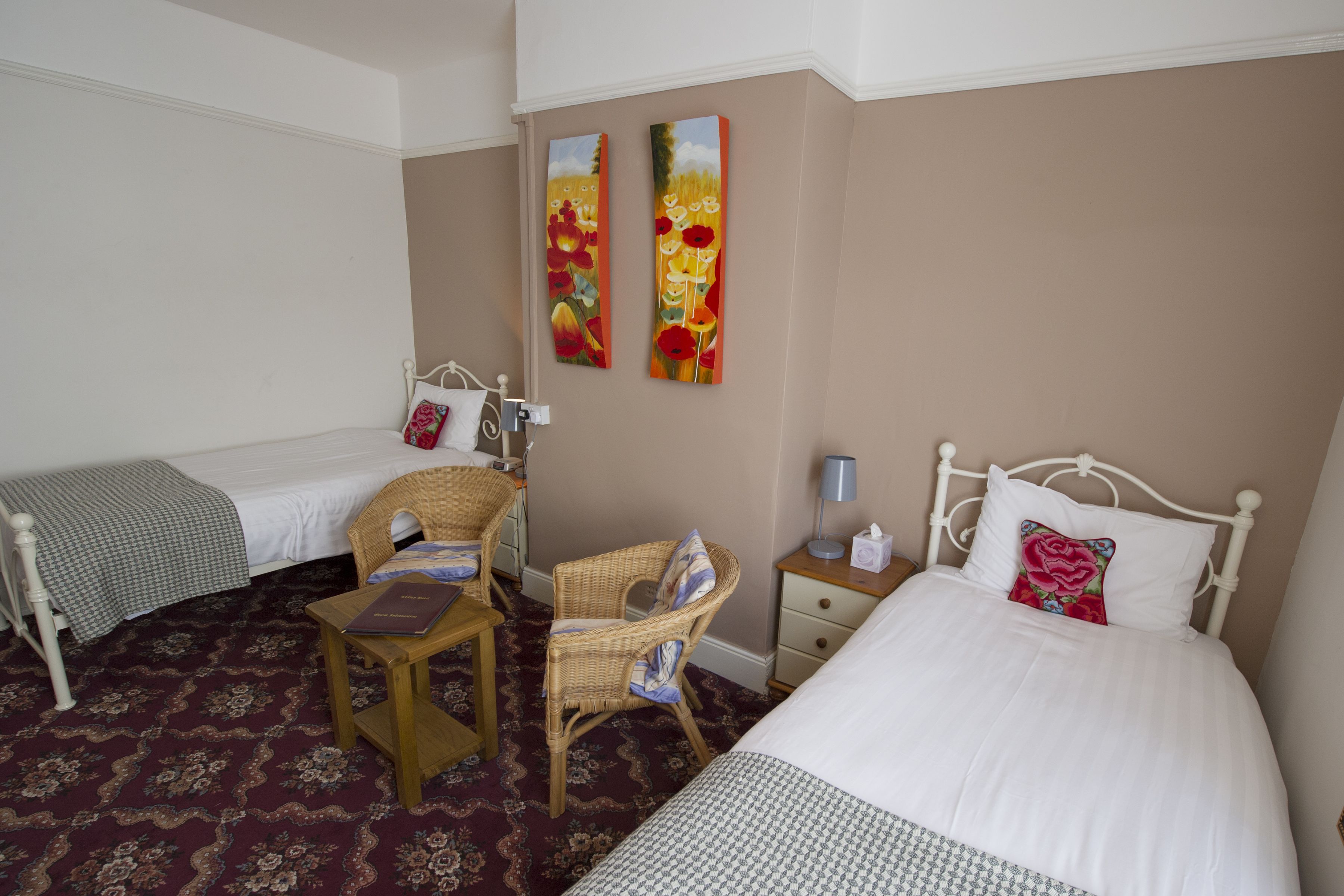A hotel room with two single beds, a small table, a chair, a nightstand, and two paintings on the wall.