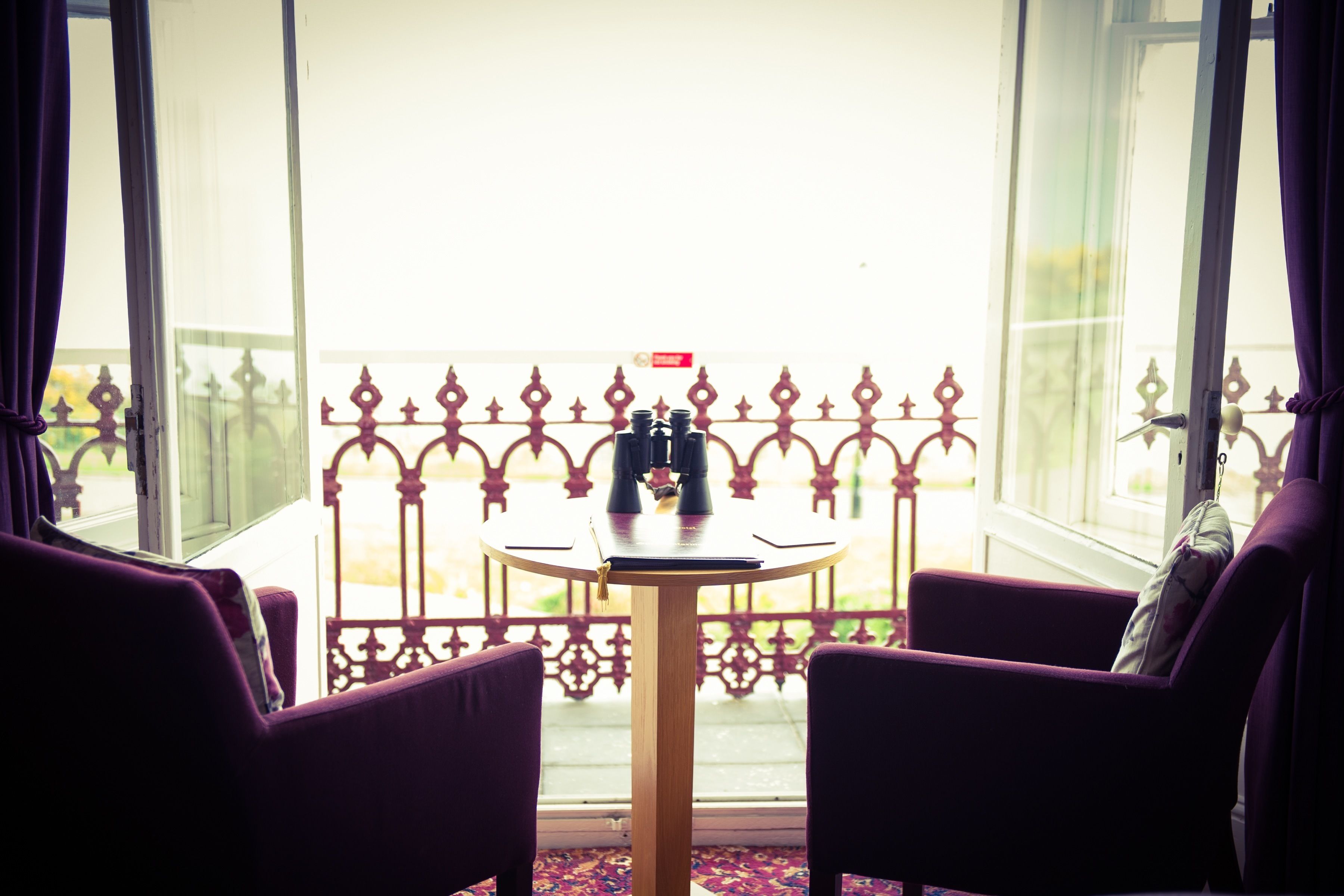 A room with a table and two chairs, a window with curtains, and a balcony with a decorative railing.