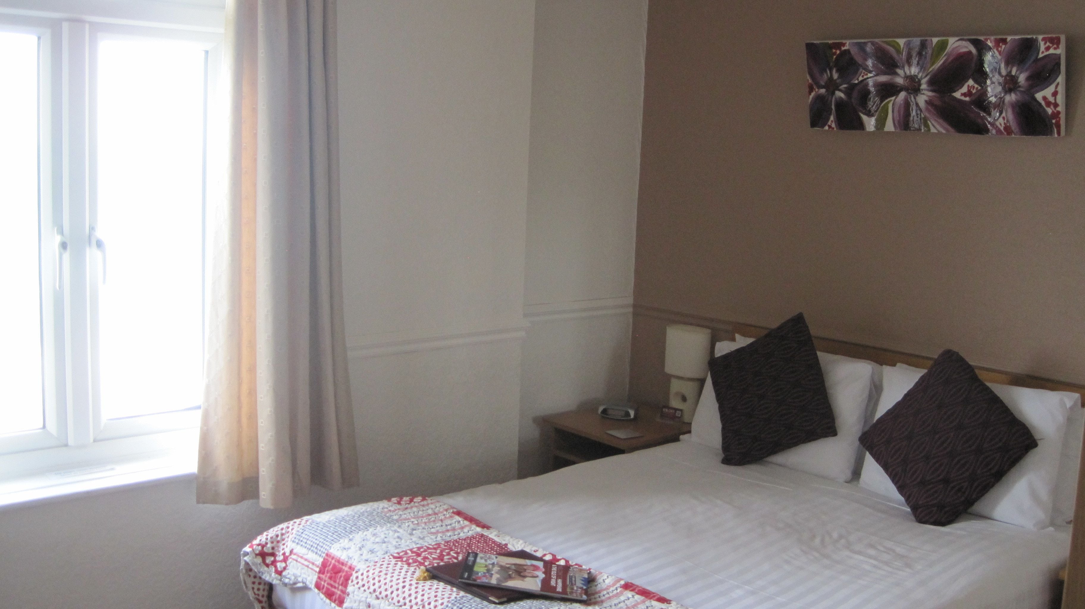Una habitación de hotel con cama, mesita de noche, un cuadro y una ventana con cortinas.