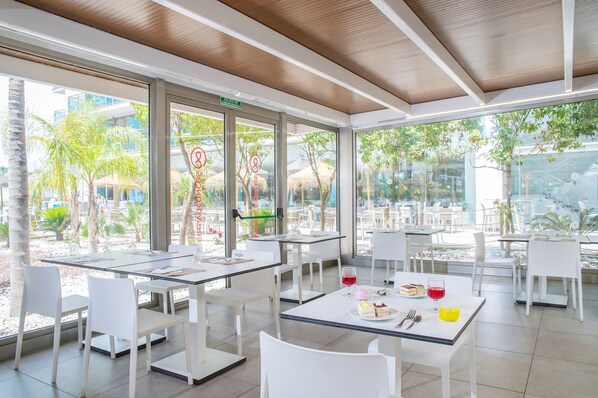 A modern outdoor dining area with glass tables and white chairs, surrounded by glass walls and a view of palm trees and buildings.