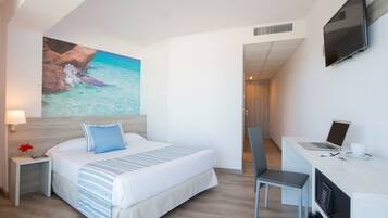 Habitación de hotel con cama, escritorio, laptop, televisor y un cuadro con una playa.