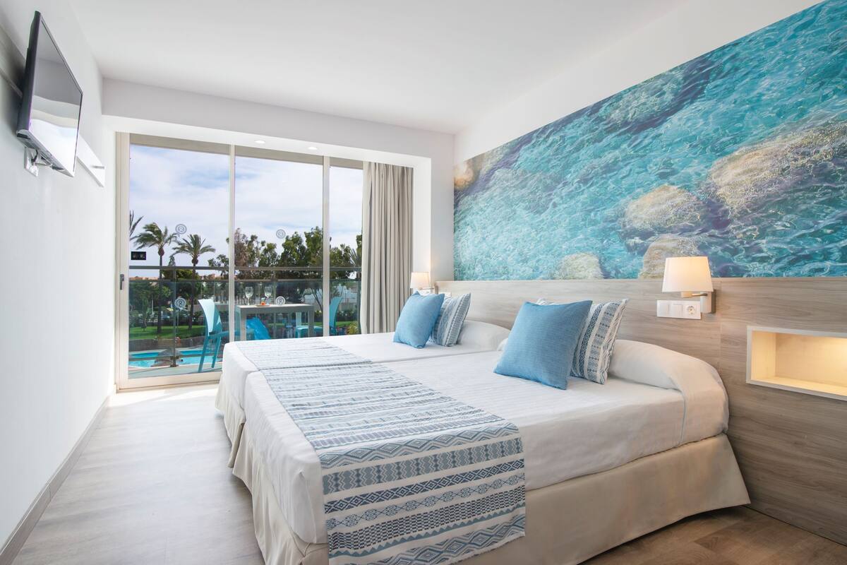 Ein Hotelzimmer mit einem großen Bett, einem Meeresbild, einem an der Wand montierten Fernseher und einem Balkon mit Blick auf Palmen.
