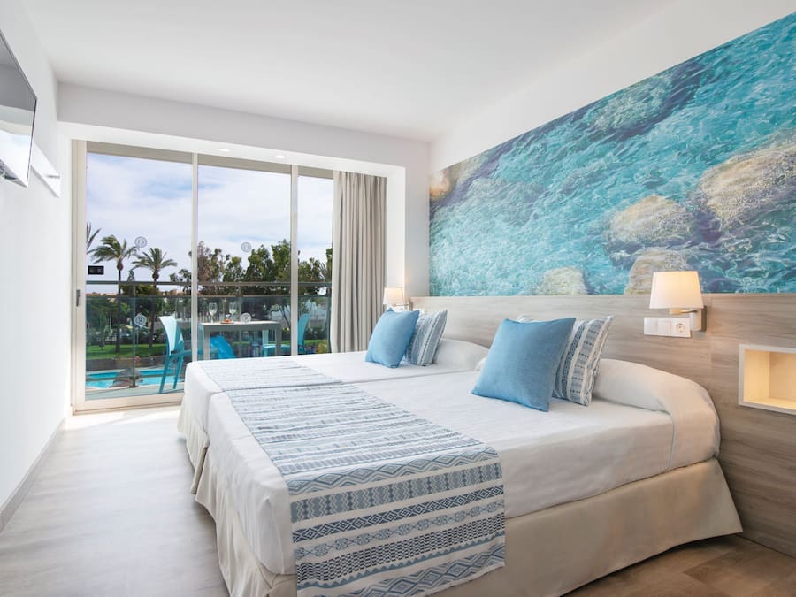 A hotel room with a large bed, a painting of the ocean, a TV mounted on the wall, and a balcony with a view of palm trees.