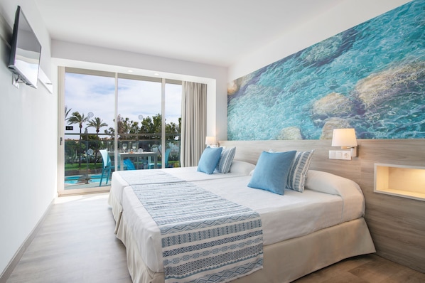 A hotel room with a large bed, a painting of the ocean, a TV mounted on the wall, and a balcony with a view of palm trees.