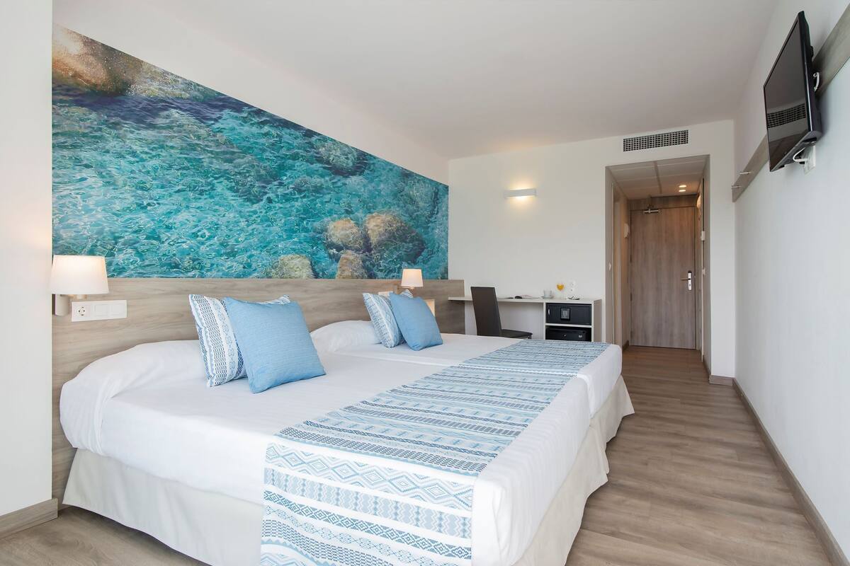 Ein Hotelzimmer mit einem großen Bett, einem Schreibtisch, einem Fernseher und einem Gemälde, das eine blaue Meereslandschaft zeigt.