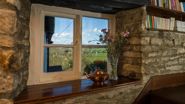 A rustic room with a stone wall, a wooden bookshelf, a window with a view of greenery, and a brass teapot on a wooden ledge. - Willance House Guest House (Richmond)