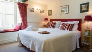 A neatly made bed with a white bedspread, red and white striped pillows, and a red headboard. There are framed pictures on the wall, a window with red curtains, and a bedside table with a lamp. - Willance House Guest House (Richmond)
