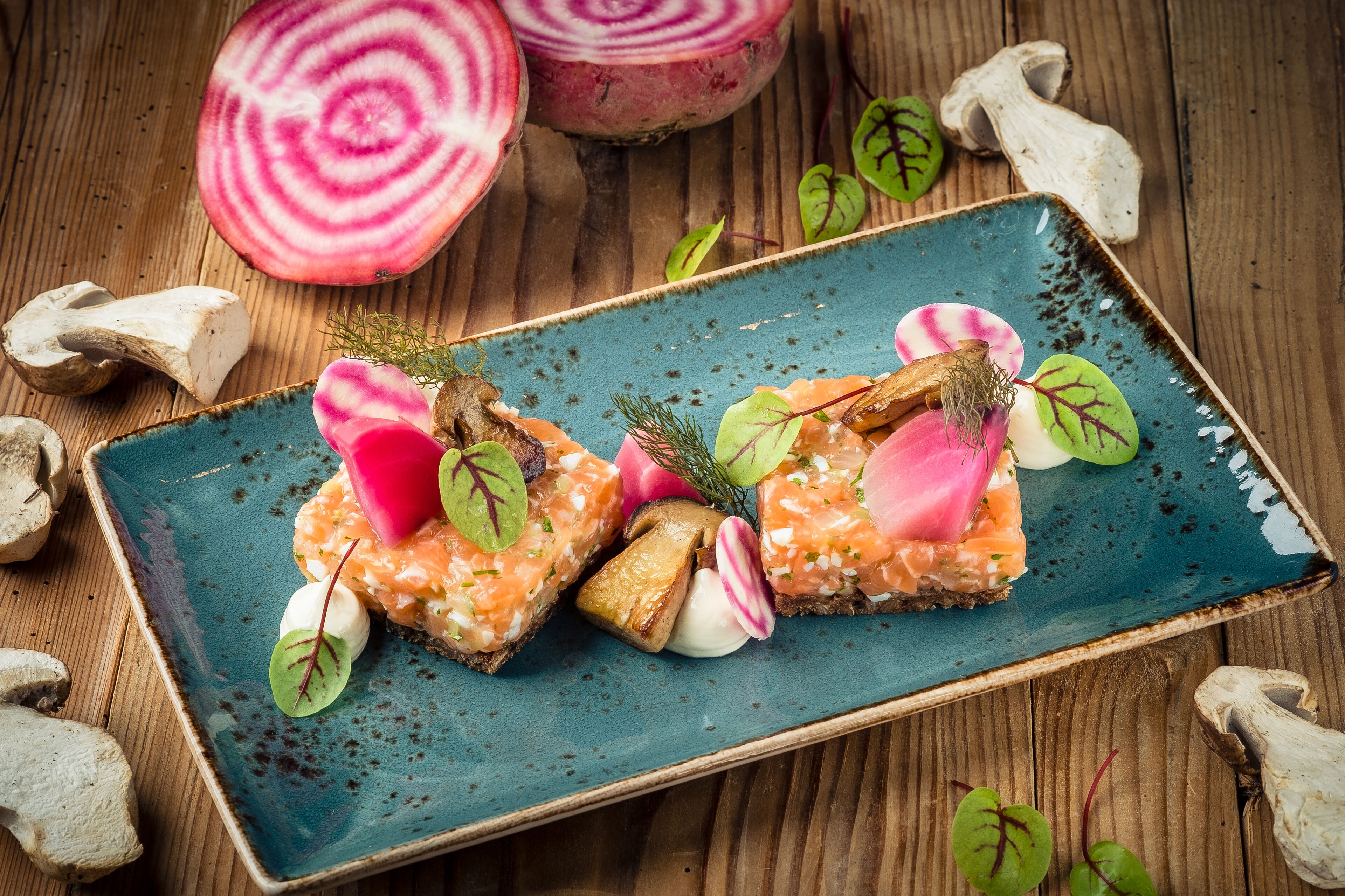 A rectangular plate with three pieces of salmon tartare on a rustic blue ceramic plate, surrounded by fresh vegetables and herbs.