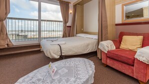 A hotel room with a bed, a red sofa, a round table with a lace tablecloth, and a large window with curtains. - Lynton Apartments (Blackpool)