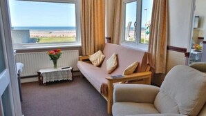 A room with a sofa, a chair, a small table with a vase, and a view of the beach through large windows. - Lynton Apartments (Blackpool)