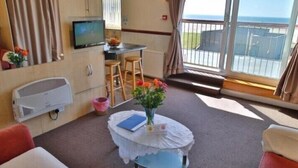 A room with a TV, a small table with a vase, a chair, and a view of the sea. - Lynton Apartments (Blackpool)