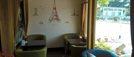 A room with a mural of the Eiffel Tower, two green chairs, a small table, and a view of Ayutthaya City Park through a window.