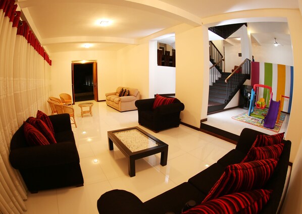 Un salon avec un canapé noir, un canapé beige et une table basse. On y trouve un escalier, et au pied de celui-ci, un espace de jeux coloré.