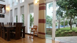 A dining area with a wooden table and chairs, a large window with a view of greenery, and a patterned curtain. - Capital Villa (Battaramulla)