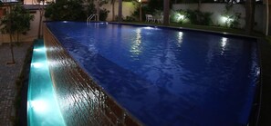 A rectangular pool with a clear blue water surface, surrounded by a low brick wall and a small garden area with trees and a white outdoor table and chairs. - Capital Villa (Battaramulla)