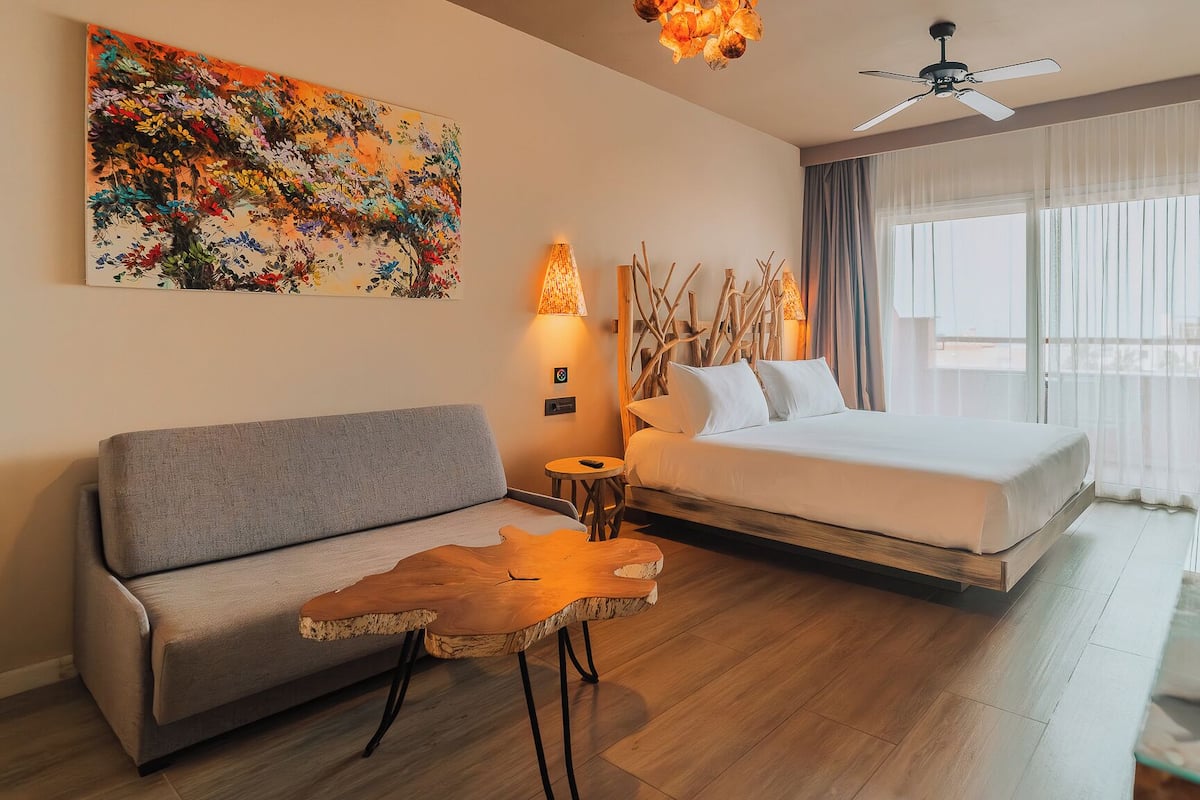 Habitación de hotel con cama, sofá, mesa de centro, un cuadro, lámpara, ventilador de techo y ventana con cortinas.