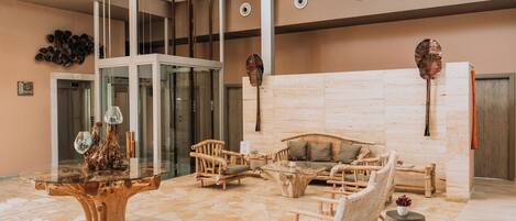 A lobby with a glass elevator, wooden furniture, a central wooden table, and a stone-tiled floor.