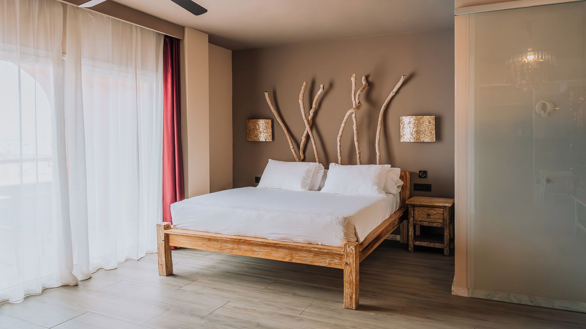 Un lit avec un cadre en bois, une table de chevet, un luminaire fixé au mur et une fenêtre avec des rideaux légers.