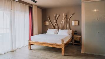 Una cama con estructura de madera, una mesita de noche, un aplique de pared y una ventana con cortinas de gasa.