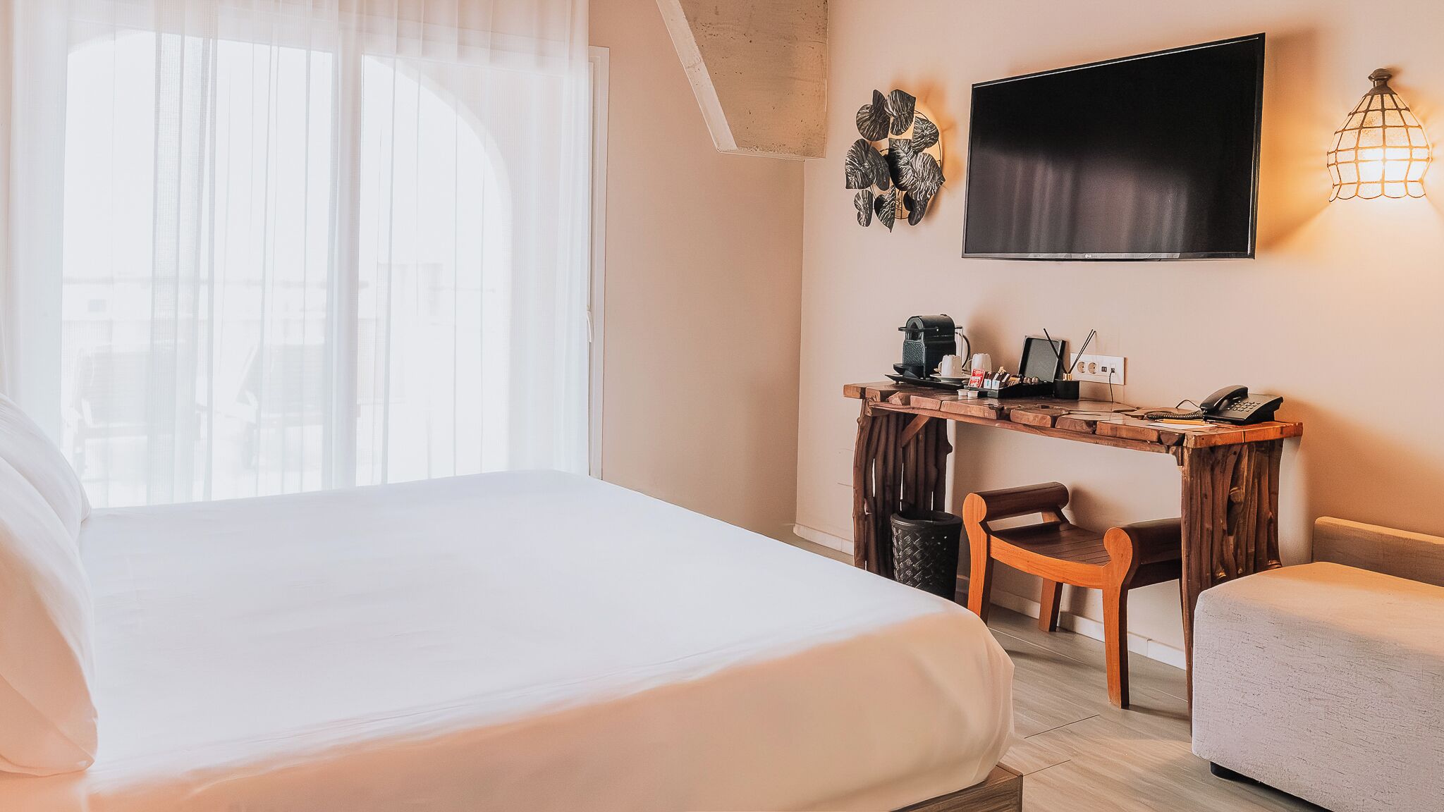 A hotel room with a bed, a desk with a chair, a TV, and a wall-mounted light fixture.