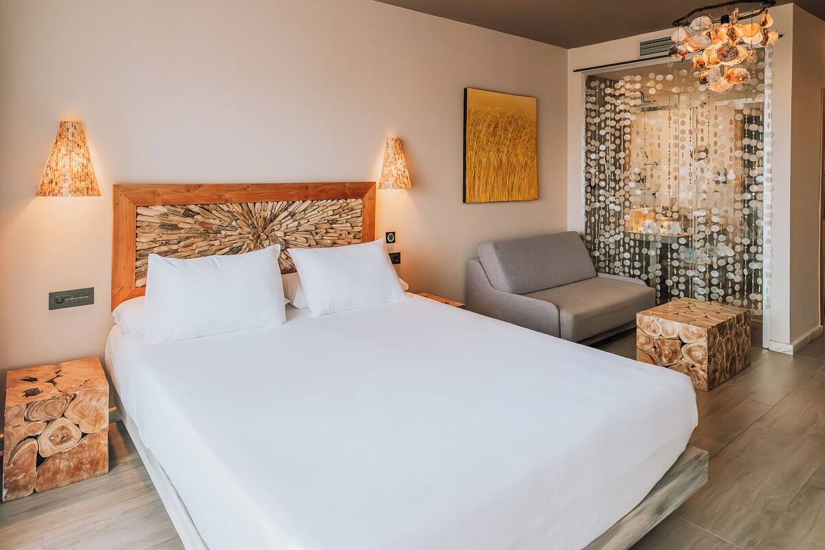 Habitación de hotel con una cama grande, un sofá gris, un cabecero de madera, dos lámparas de pared y un adorno en la pared.