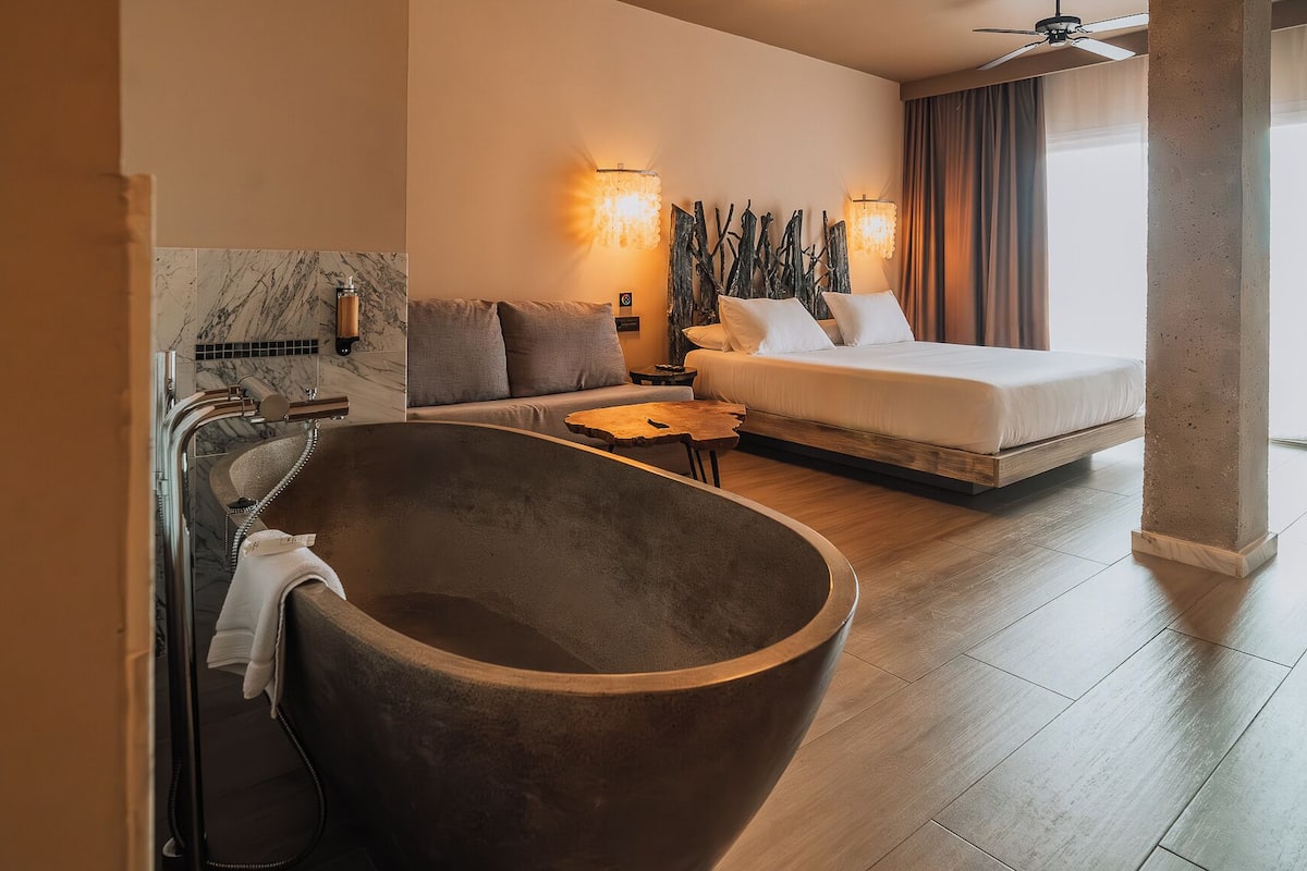 Una habitación de hotel moderna con una bañera independiente grande, una cama con ropa blanca, un sofá con cojines, una mesita de madera pequeña y una lámpara fijada a la pared.
