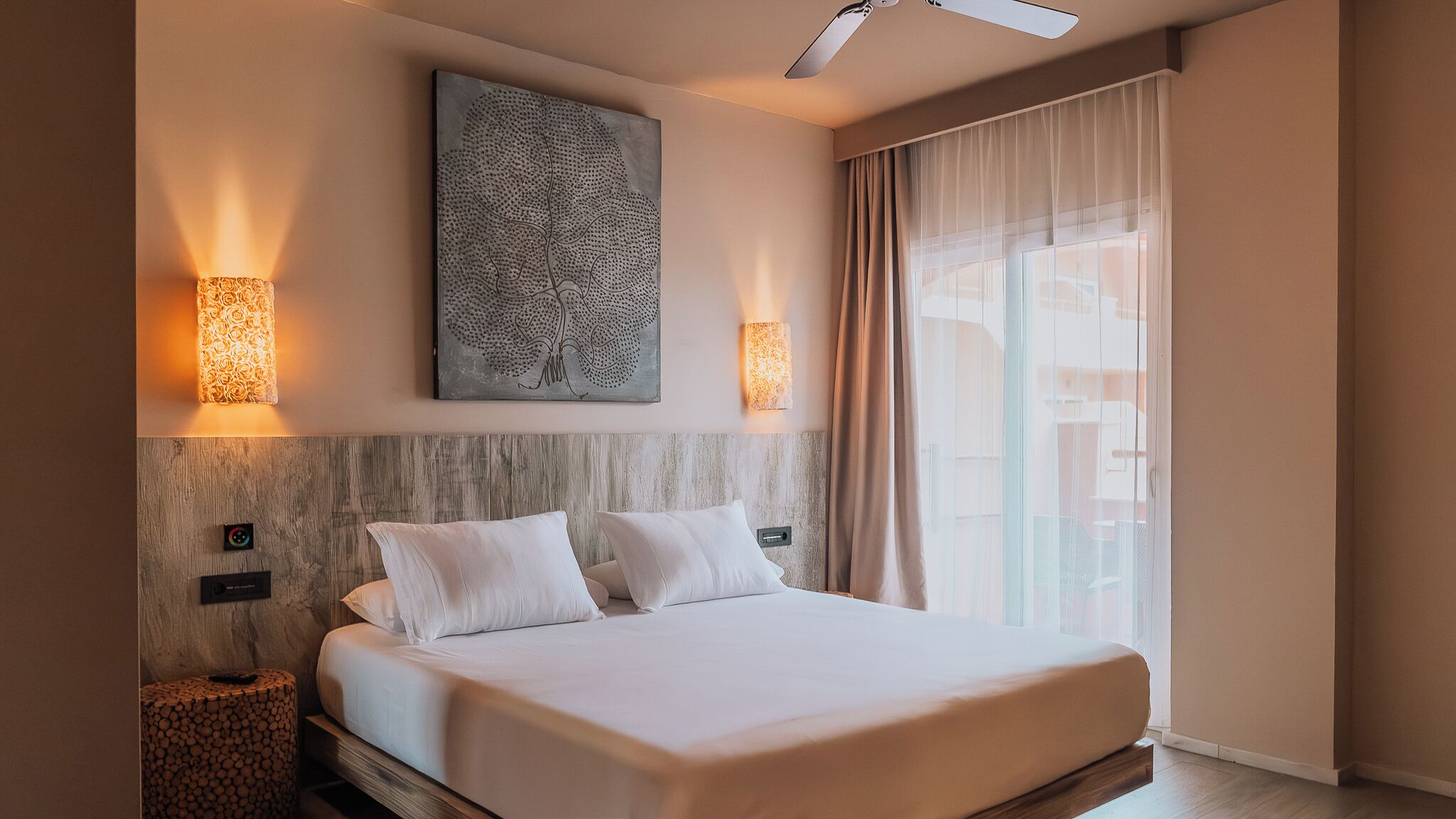A hotel room with a bed, a wall-mounted artwork, two wall sconces, a ceiling fan, and a large window with curtains.