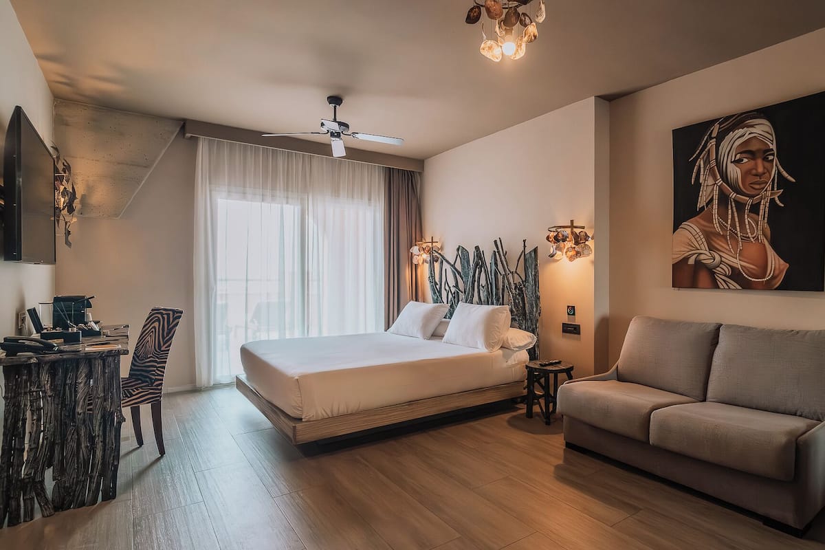 Una habitación de hotel moderna con cama, sofá, escritorio con computadora y un cuadro grande en la pared.