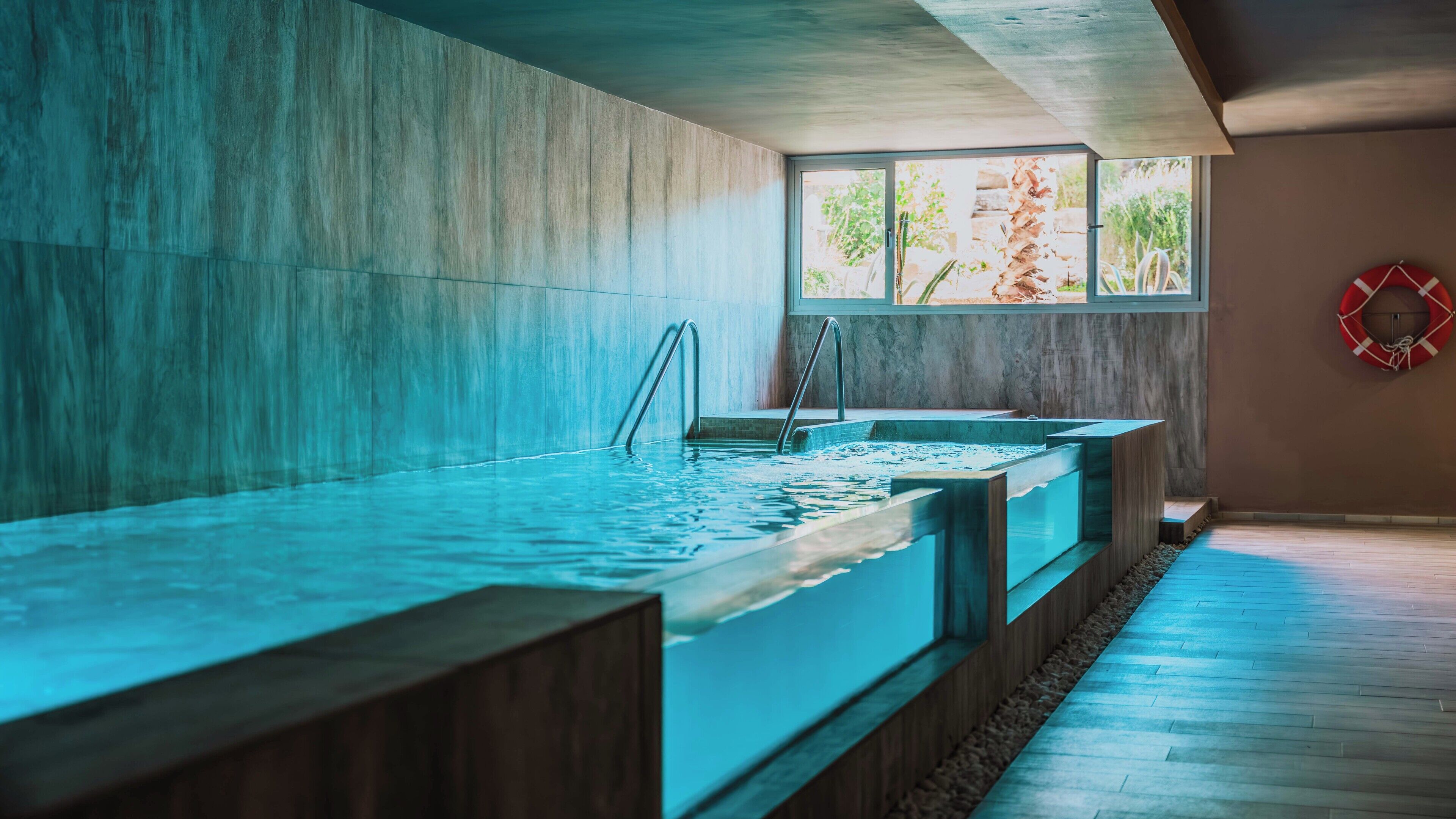 A modern indoor pool with clear water, wooden steps, and a view of greenery through a window.