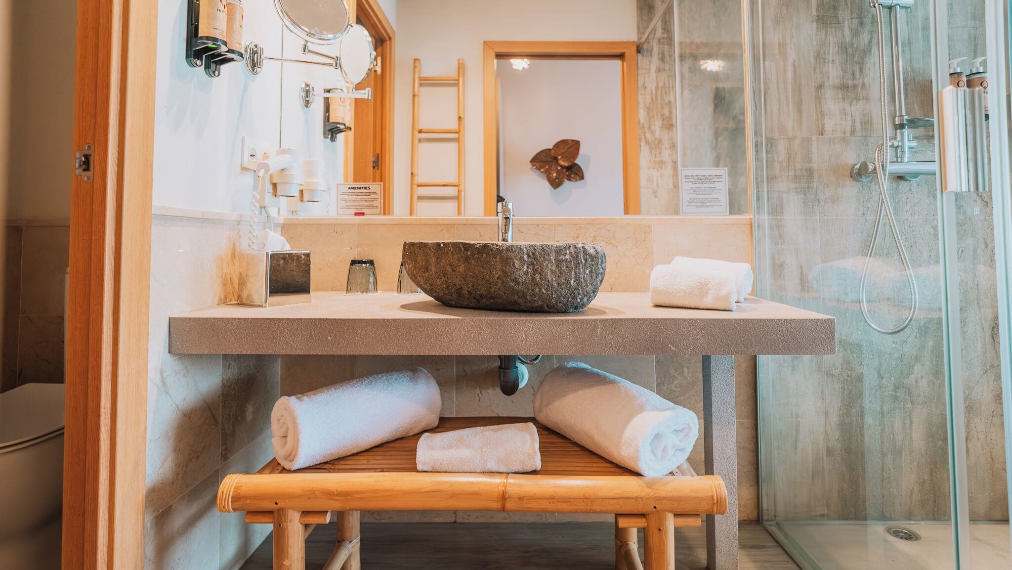 Un baño con lavamanos de piedra, una banca de madera con toallas, una cabina de ducha de vidrio y una puerta de madera.