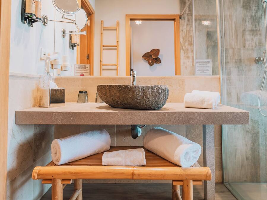 Un baño con lavamanos de piedra, una banca de madera con toallas, una cabina de ducha de vidrio y una puerta de madera.